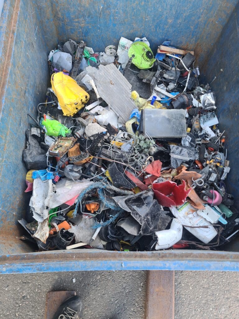 En blå affaldscontainer fyldt med forskellige typer knust plast, metaldele og elektronisk affald klar til sortering og genanvendelse.