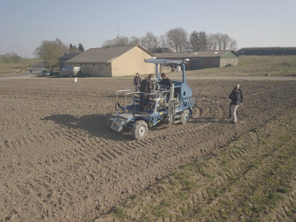 Blå selvkørende autonom landbrugsmaskine prototype under fieldtest med ingeniører på en mark.