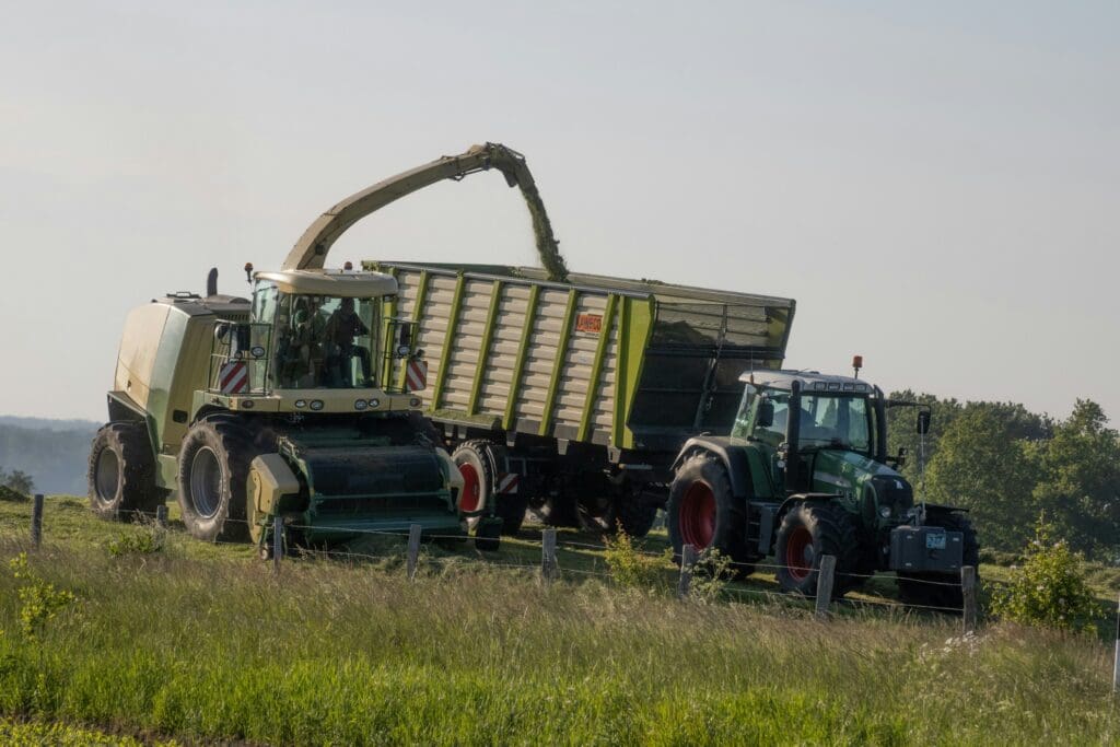 En selvkørende finsnitter og en traktor med vogn i gang med græshøst på en mark, repræsenterende avanceret landbrugsteknologi.