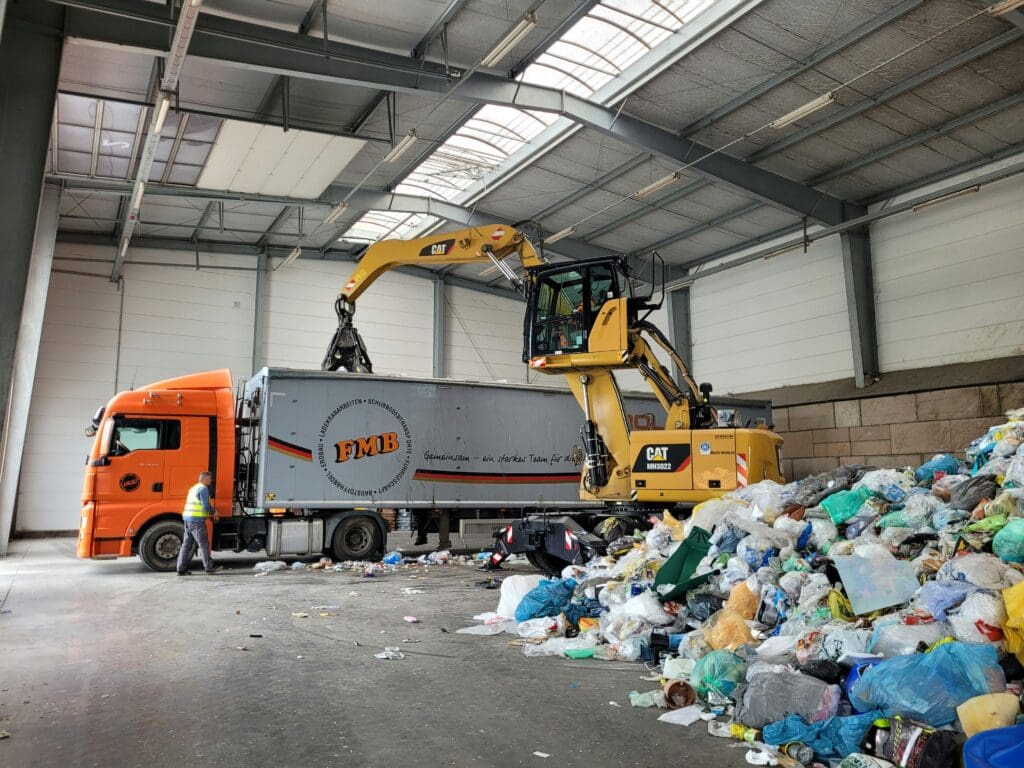 En gul CAT MH3022 materialehåndteringsmaskine læsser sorteret plastikaffald i en lastbil i en genanvendelseshal.