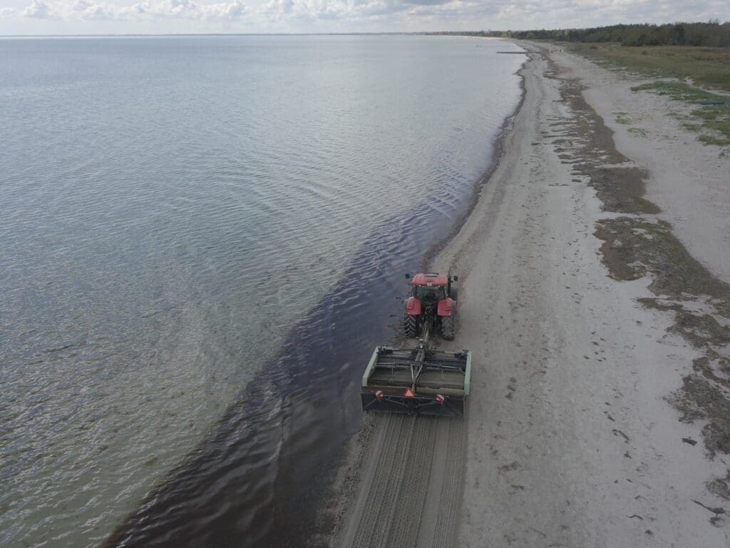 En rød traktor trækker en specialudviklet strandrensningsmaskine langs en kystlinje ved hav og fjord.