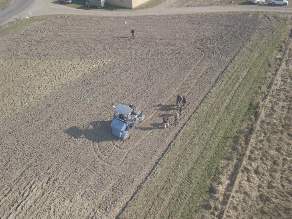 Luftfoto af en blå selvkørende landbrugsmaskine under fieldtest på en pløjet mark med ingeniører fra Phidan Engineering.