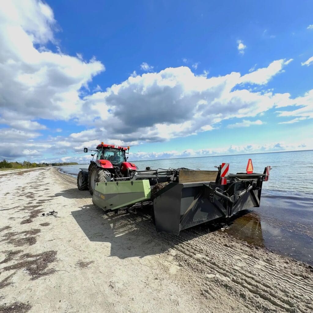 En rød traktor trækker en avanceret Root Up 4000 strandrenser udviklet af Phidan Engineering på en sandstrand.