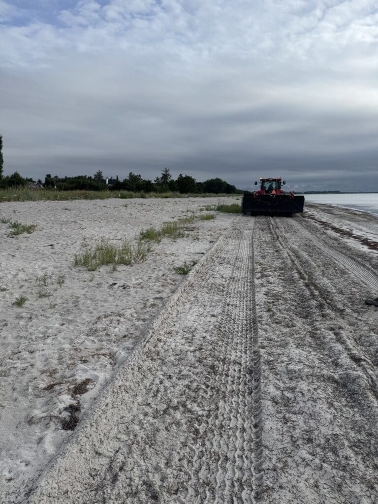 En rød traktor med en specialiseret strandrenser kører på en hvid sandstrand ved havet.