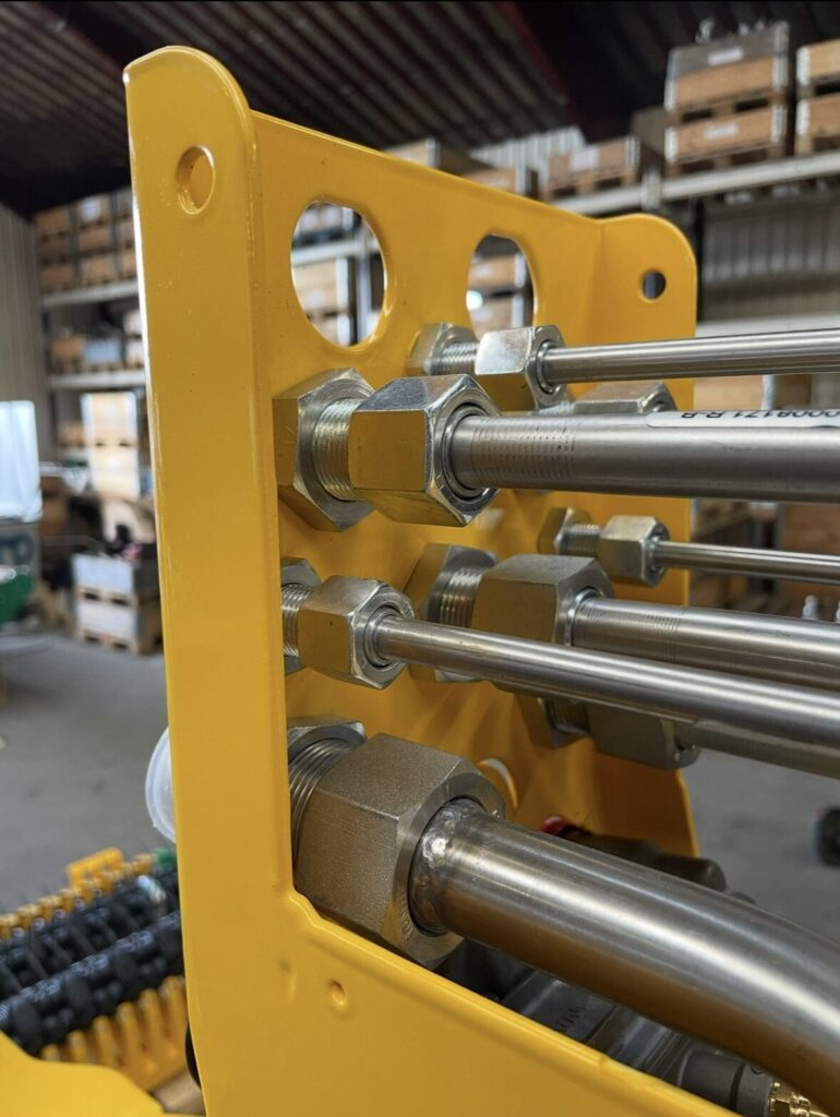 Nærbillede af hydrauliske rør og bulkhead-forskruninger monteret i et gult metalbeslag på en maskine.