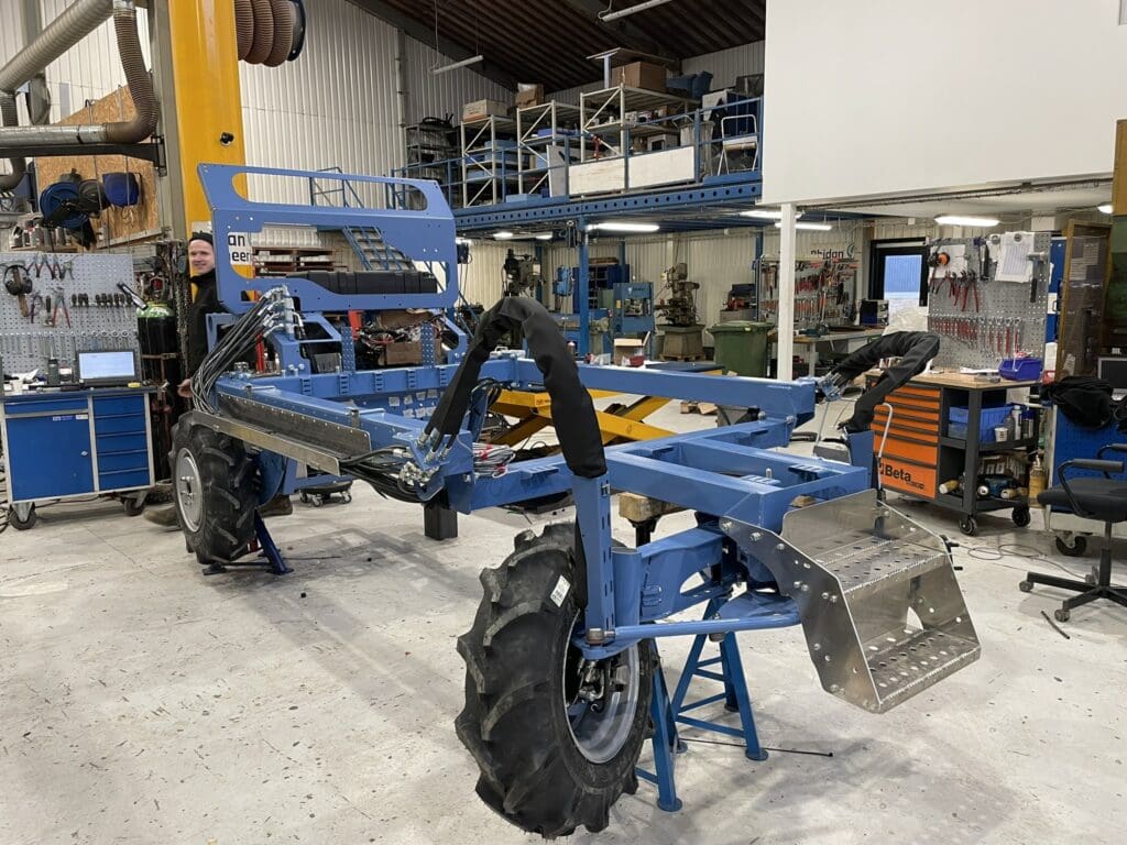 Blå maskinkonstruktion til en selvkørende maskine med store terrængående dæk og synlig hydraulik under montering i et værksted.