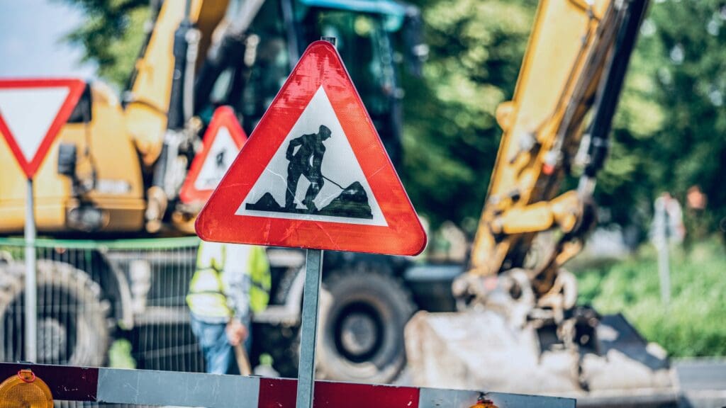 Vejarbejdsskilt foran en gul gravemaskine, der illustrerer produktudvikling og teknisk dokumentation for entreprenørmaskiner hos Phidan Engineering.