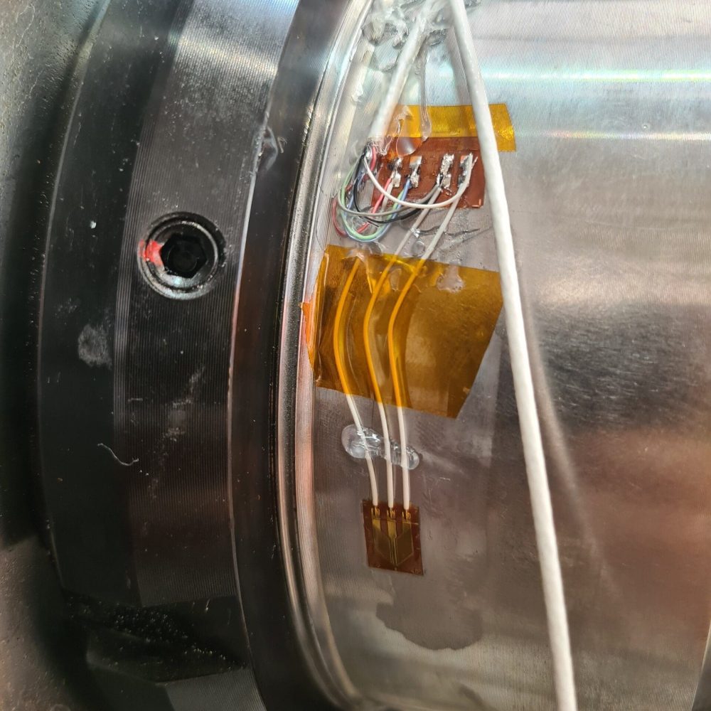 Nærbillede af strain gauge monteret på en cylindrisk metalkomponent med ledninger og beskyttelsestape til datalogning.